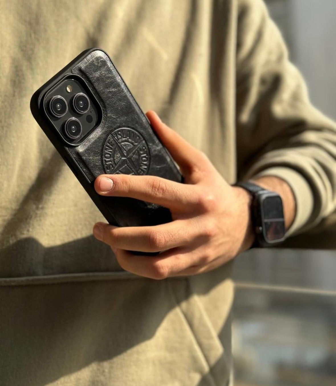 Man holding iPhone with black Stone Island case, wearing beige sweatshirt and smartwatch