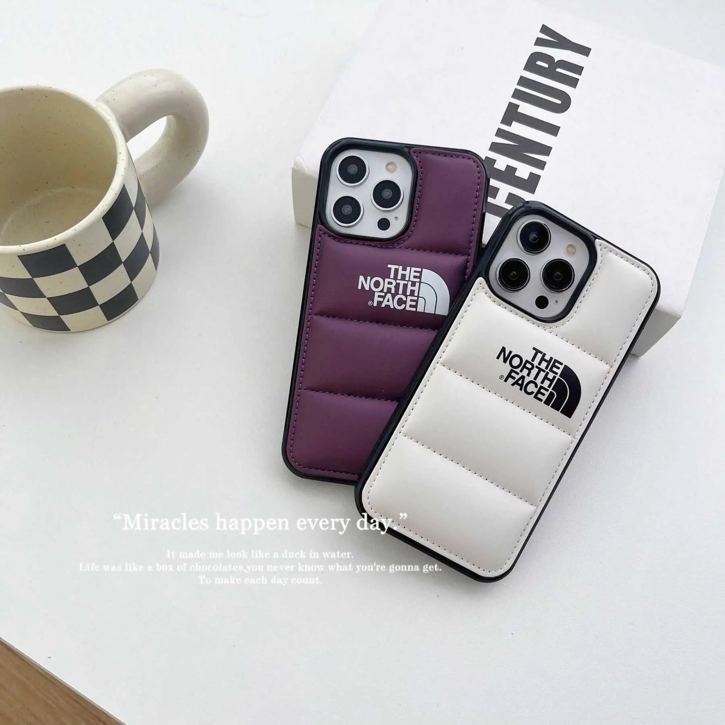 The North Face puffer iPhone cases in purple and white on table with checker mug and book