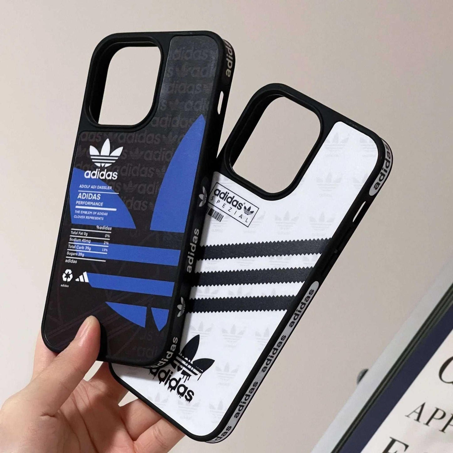 Hand holding two Adidas iPhone cases with logo and stripe designs, black and white backgrounds