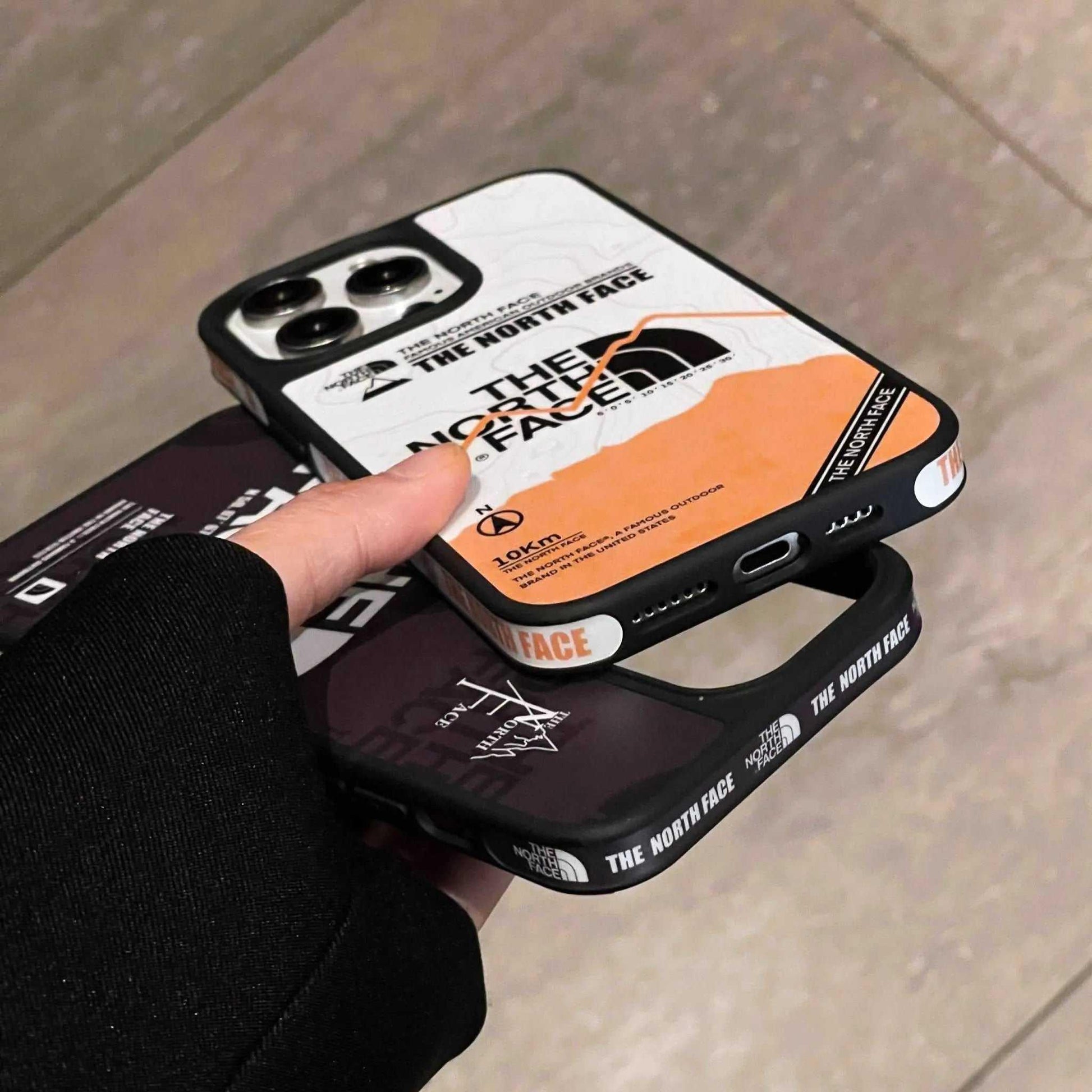 Hand holding two The North Face branded iPhone cases with bold logo designs