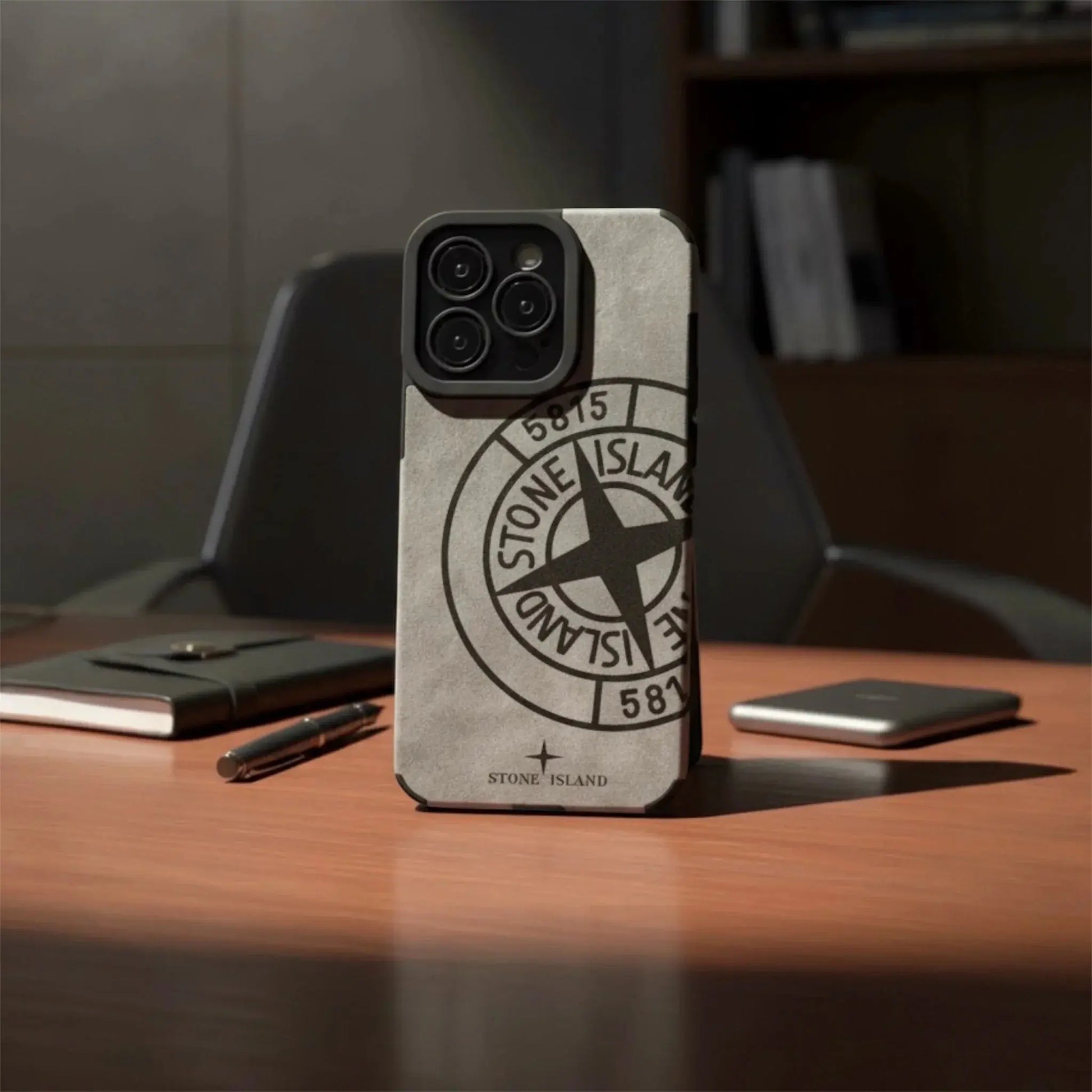 Stone Island logo iPhone case on office desk with notebook, pen, and chair in background