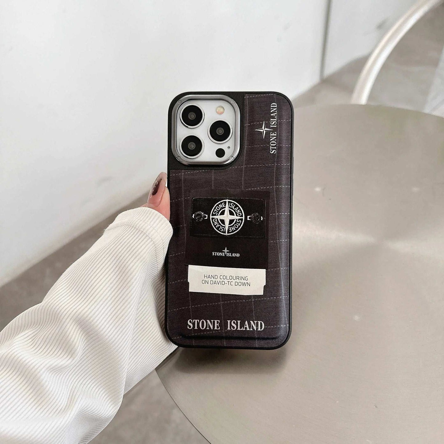 Stone Island iPhone case with logo and patch design held in hand on a gray table.