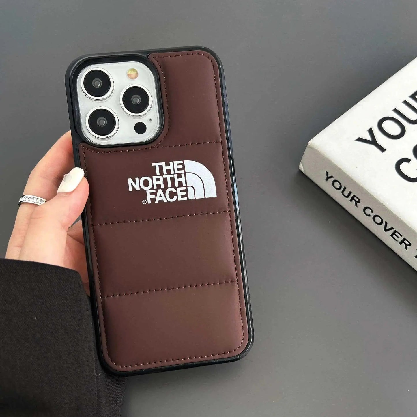 Brown padded iPhone case with The North Face logo, hand holding phone on grey background